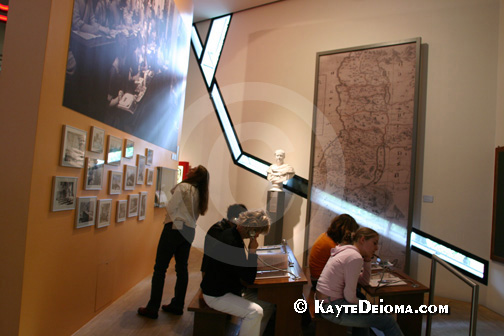 A streak of natural light comes through a slash of window into an exhibit area where a woman looks at a wall display as other seated visitors listen to an audio program while viewing visual materials at the Jewish Museum, Berlin, Germany.